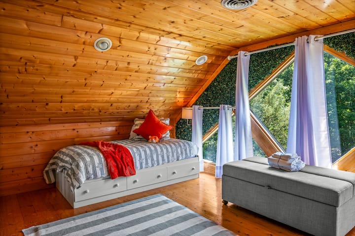 Udoban prostor u potkrovlju s kosim drvenim stropom, dvostrukim krevetom i velikim trokutastim prozorima koji propuštaju prirodno svjetlo. Nježni sivi tonovi, prugasti tepih i zeleni detalji na zidu stvaraju topao, moderan planinski ugođaj.