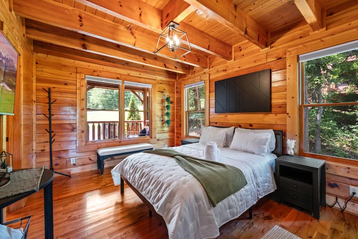 Ugodna spavaća soba s drvenim oblogama i zasvođenim stropom. Udoban krevet s bijelom posteljinom i zelenim pokrivačem okrenut je prema televizoru koji je postavljen na zid. Veliki prozori pružaju pogled na šumu i stvaraju mirno utočište u planinama Smoky Mountains.
