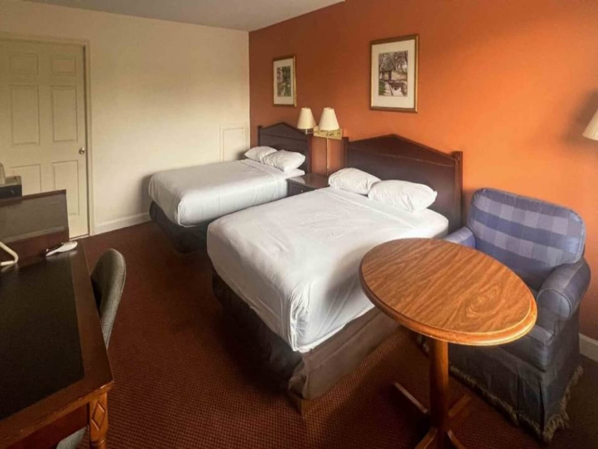 The room features two full beds with white linens arranged neatly. A small wooden table accompanied by a blue upholstered chair is positioned beside the beds. A work desk is located against the wall, with a lamp providing light. Soft orange accents adorn the wall.