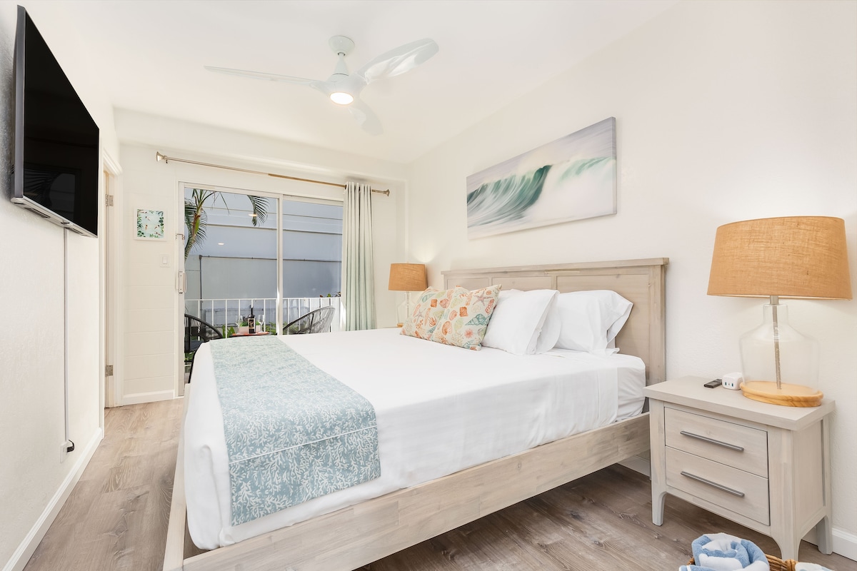 A bright bedroom is featuring a spacious bed adorned with light bedding and decorative pillows. Two lamps provide soft lighting beside the bed, while a wall-mounted television is positioned across from it. A large window allows natural light to enter, framed by sheer curtains.
