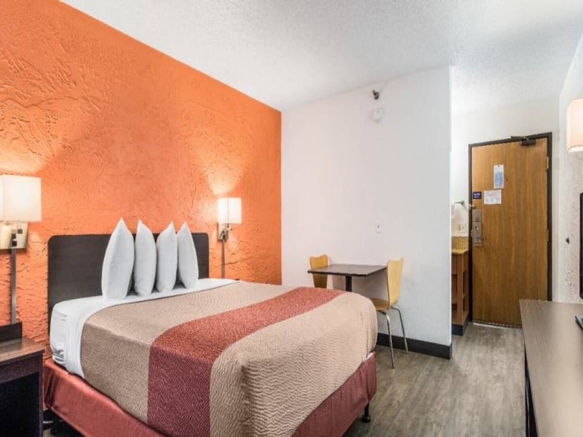 A comfortable guest room features a king bed with plush pillows. An accent wall in orange adds warmth to the space. A small table with two chairs is positioned in the corner, while a door leads to the room’s entrance.