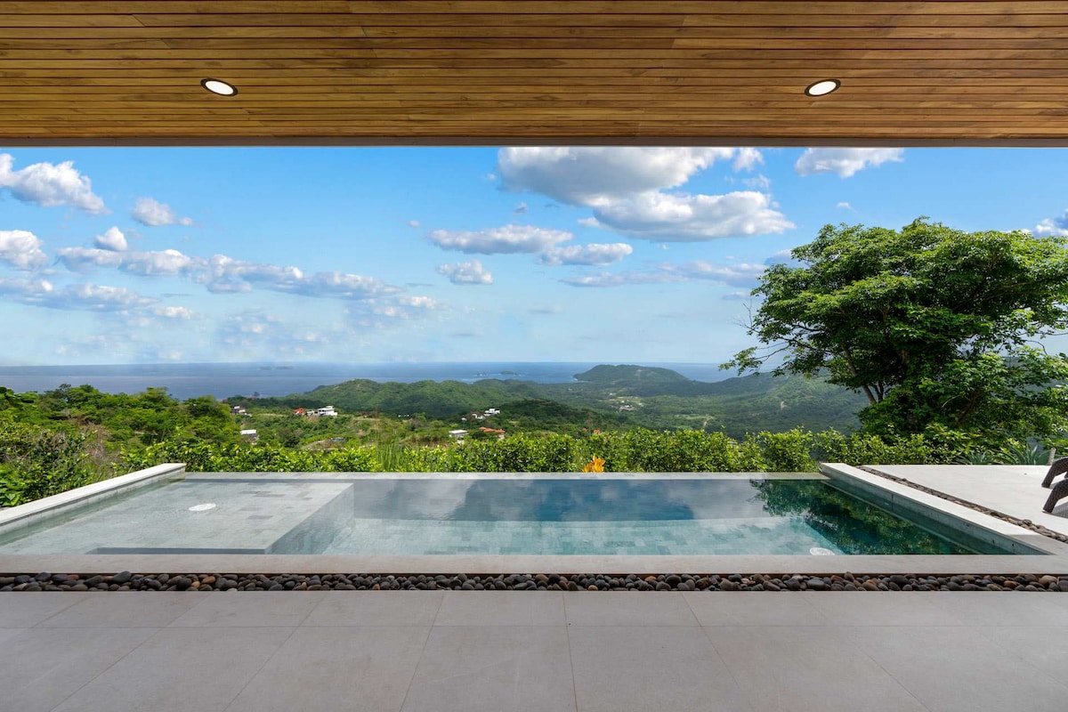 A private infinity pool is set against a backdrop of lush green hills and the expansive Pacific Ocean. The view features a clear blue sky dotted with clouds, enhancing the tranquil atmosphere of the outdoor space.