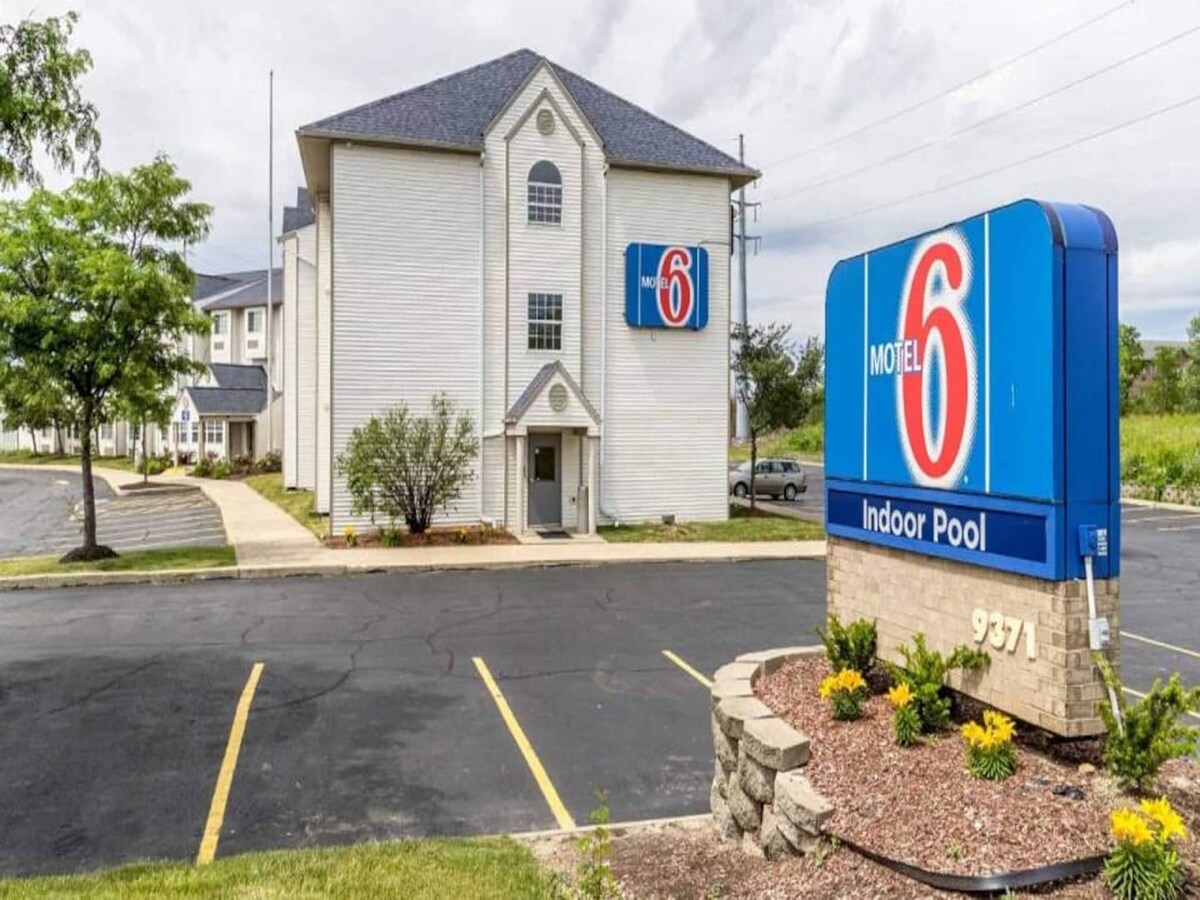 The exterior of Motel 6 features a two-story building with a prominent sign indicating an indoor pool. Neatly arranged parking spaces and low-maintenance landscaping with small shrubs and flowers frame the entrance, providing a clean and welcoming appearance.