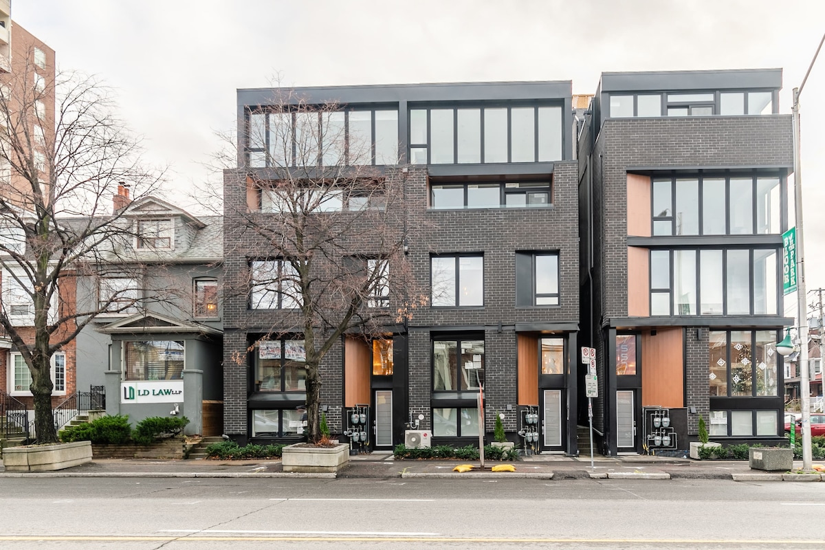 The exterior of a modern multi-unit building is displayed, featuring a sleek design with large windows and contrasting dark brick. Adjacent to the building, a traditional structure adds character to the streetscape. The sidewalk is lined with small shrubs and trees, enhancing the urban environment.
