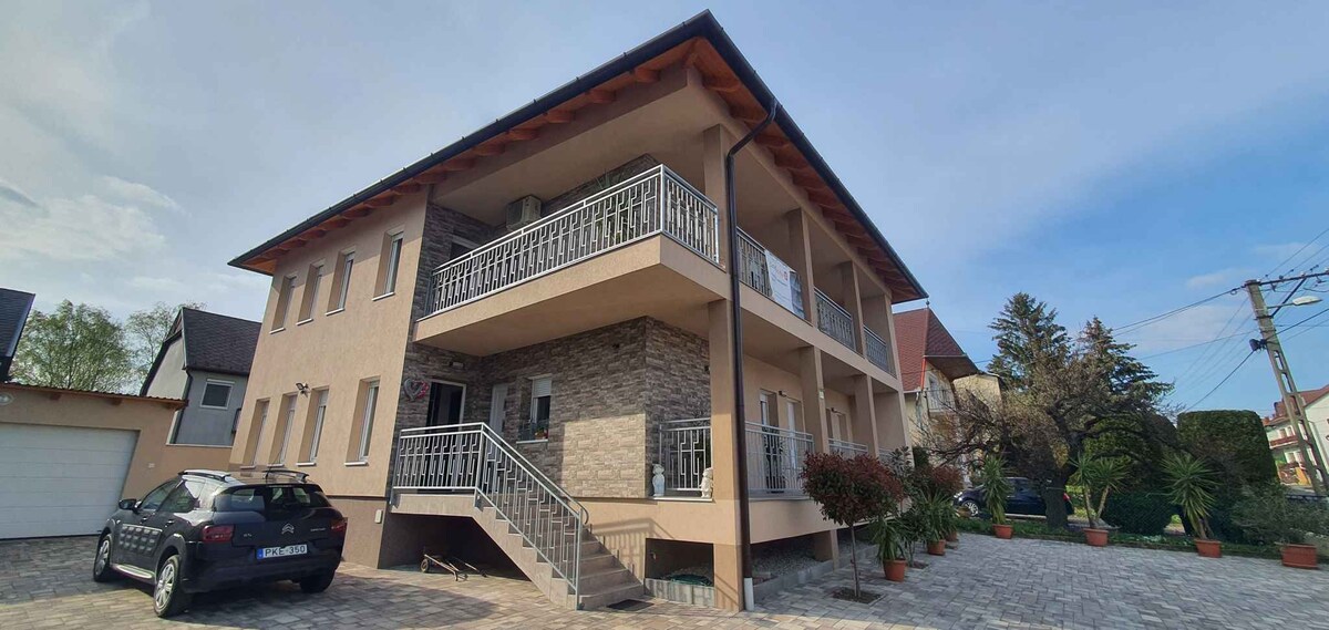 The exterior of a modern two-story building reveals a covered entrance with a staircase leading to a balcony. Potted plants are positioned along the ground level, and a parking area accommodates one vehicle. The building is surrounded by landscaped greenery and a clear blue sky.