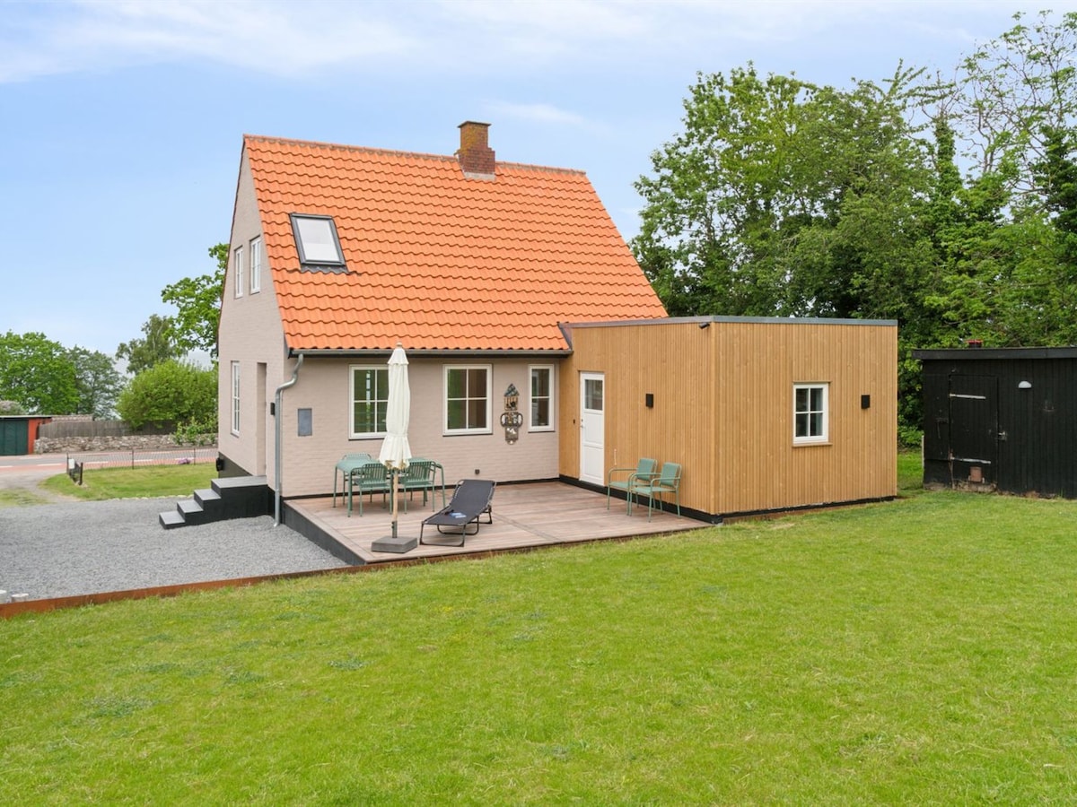 A two-story house with a red-tiled roof is surrounded by a spacious grassy area. A wooden deck features outdoor seating, including lounge chairs and a table with an umbrella. The natural landscape includes trees and greenery, enhancing the setting near the sea.