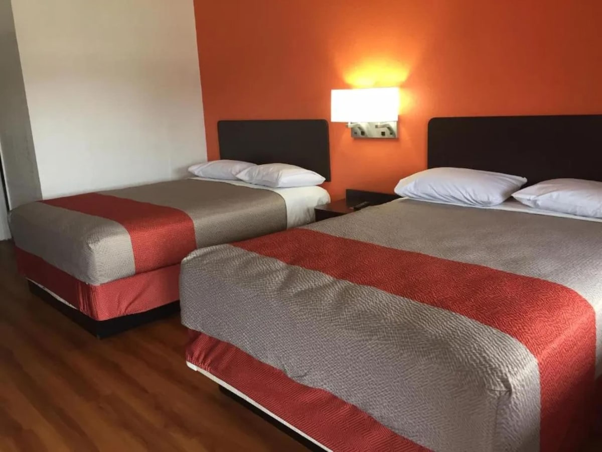 A well-lit room features two spacious beds with light-colored linens and contrasting orange bedspreads. Soft lighting from a wall-mounted fixture enhances the room's atmosphere. Wooden flooring adds warmth, while a neutral wall with an orange accent provides a pop of color.