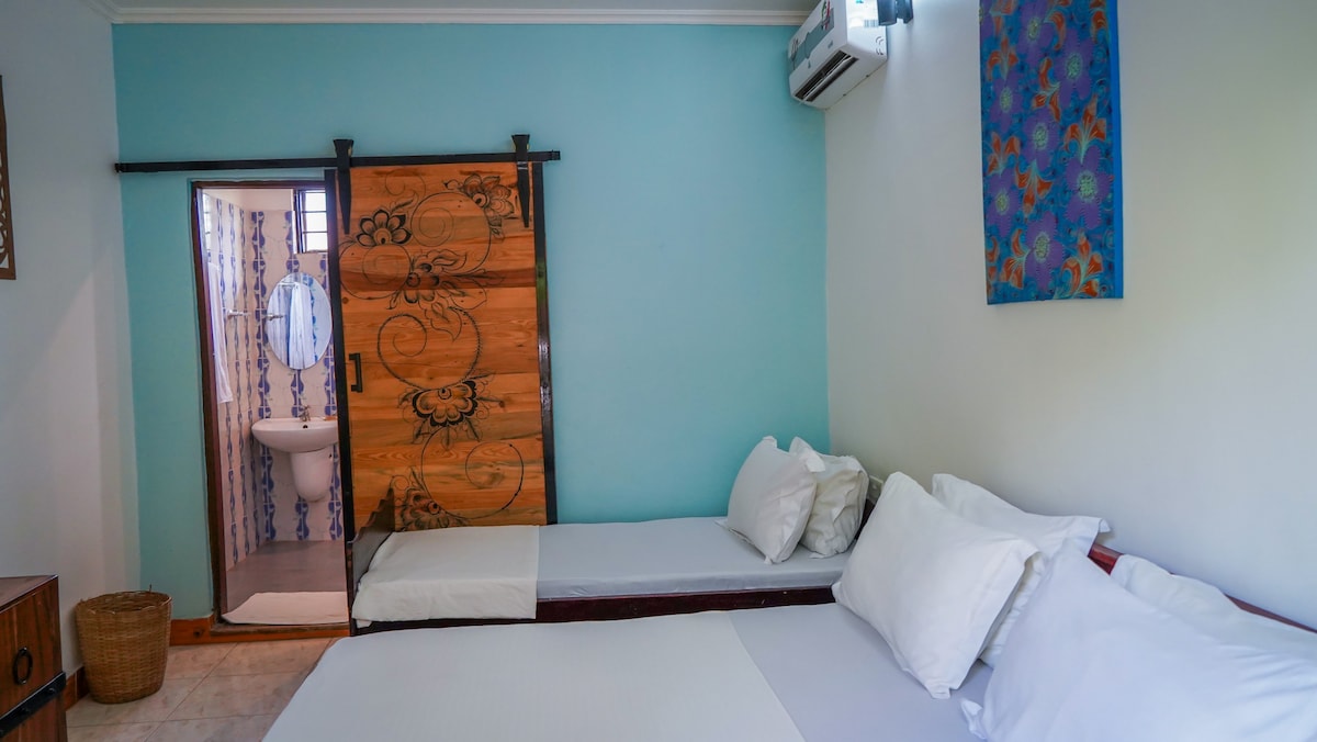 A room is presented with a queen-size bed and an additional single-size bed. The walls are painted in soft blue, and a wooden sliding door leads to a private bathroom. Air conditioning is visible, and decorative artwork adds a splash of color.