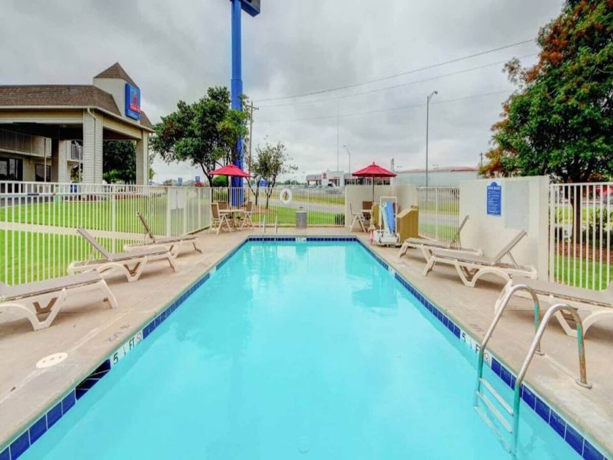 An outdoor swimming pool is showcased, surrounded by sun loungers. Umbrellas offer shade over a few seating areas. Lush green grass is visible beyond the fence that encloses the pool area, contributing to a tranquil environment.