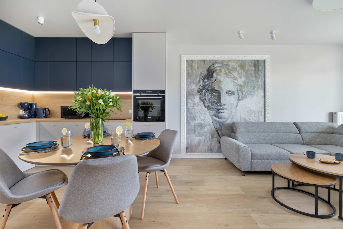 The modern kitchen area features a round dining table set for four with decorative plates and glasses. A large sofa in a light grey fabric occupies the living space, complemented by wooden coffee tables. The wall showcases an artistic mural, enhancing the aesthetic appeal.