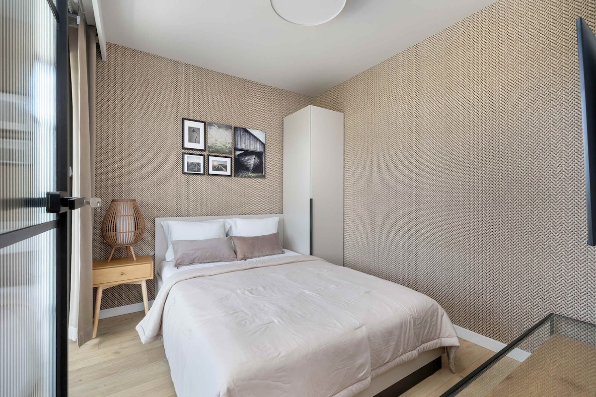 Уютна спалня в светлобежови тонове, с меко табла на леглото и произведения на изкуството, окачени на стената над леглото.