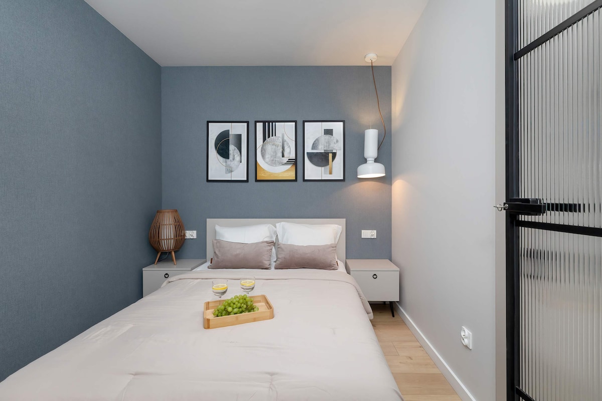 Чиста, минималистична спалня със синьо-сива стена, бели чаршафи и три рамкирани произведения на изкуството над леглото.