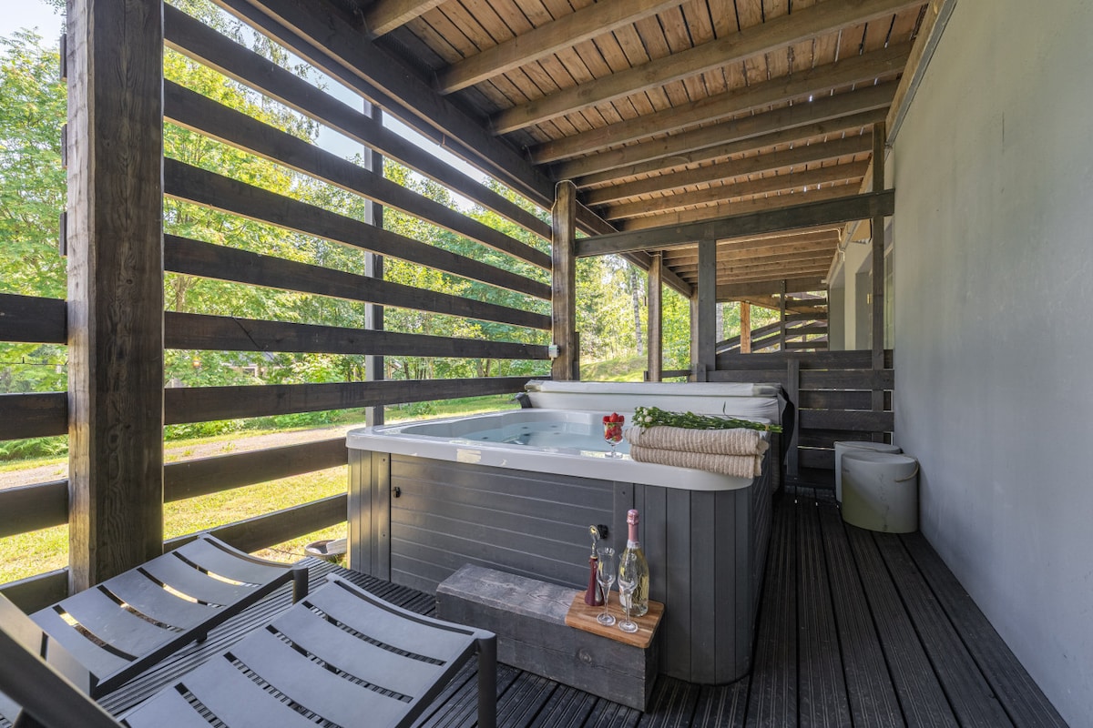 A private outdoor area features a hot tub surrounded by wooden decking and robust slatted fencing. A small table holds drinks and flowers, with lounge chairs positioned nearby for relaxation. Lush greenery is visible in the background, enhancing the serene atmosphere.