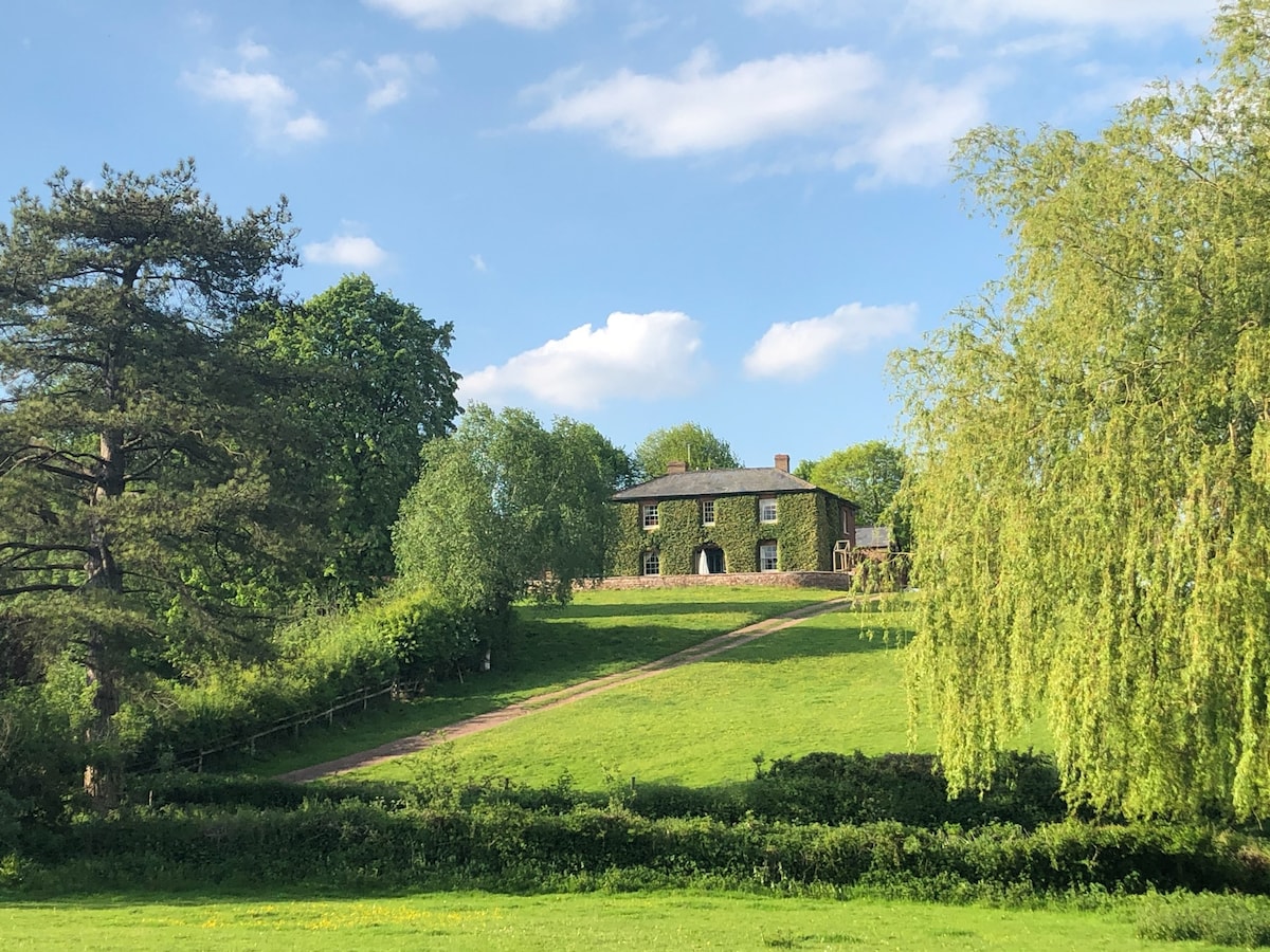 A Georgian farmhouse is positioned on a gently sloping hill, surrounded by lush greenery. The building features multiple large windows and an inviting entrance, while trees and a vibrant landscape add to the serene rural setting.