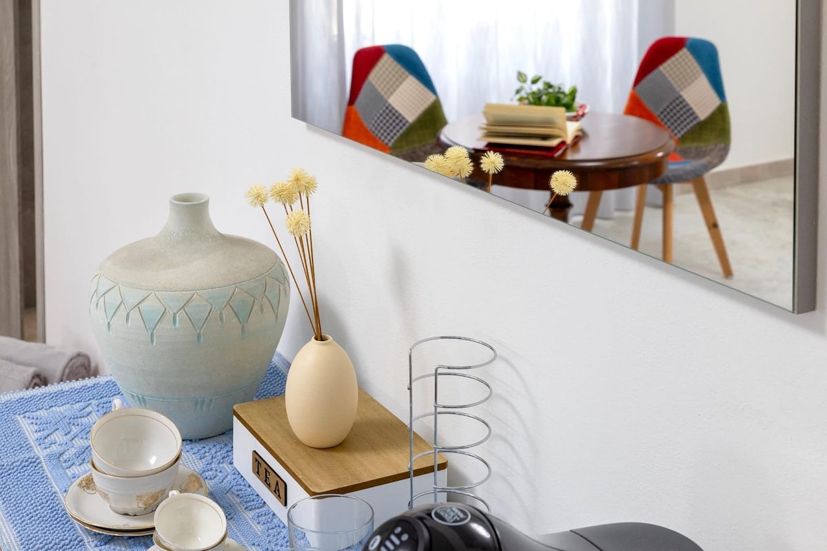 A cozy dining area is visible, featuring a round wooden table surrounded by two colorful chairs. A decorative vase with dried flowers sits on the table, while a ceramic pitcher and coffee maker are positioned nearby. A mirror reflects the arrangement, enhancing the space's openness.