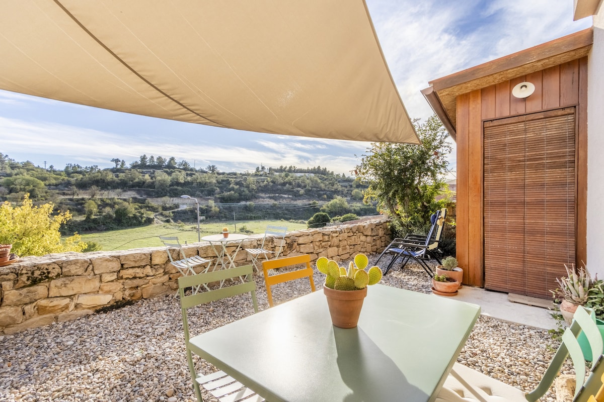 An outdoor terrace is depicted featuring a table with four colorful chairs, surrounded by gravel. A potted cactus sits at the center of the table. In the background, lush greenery and rolling hills are visible under a clear sky, with a shade awning providing protection from the sun.