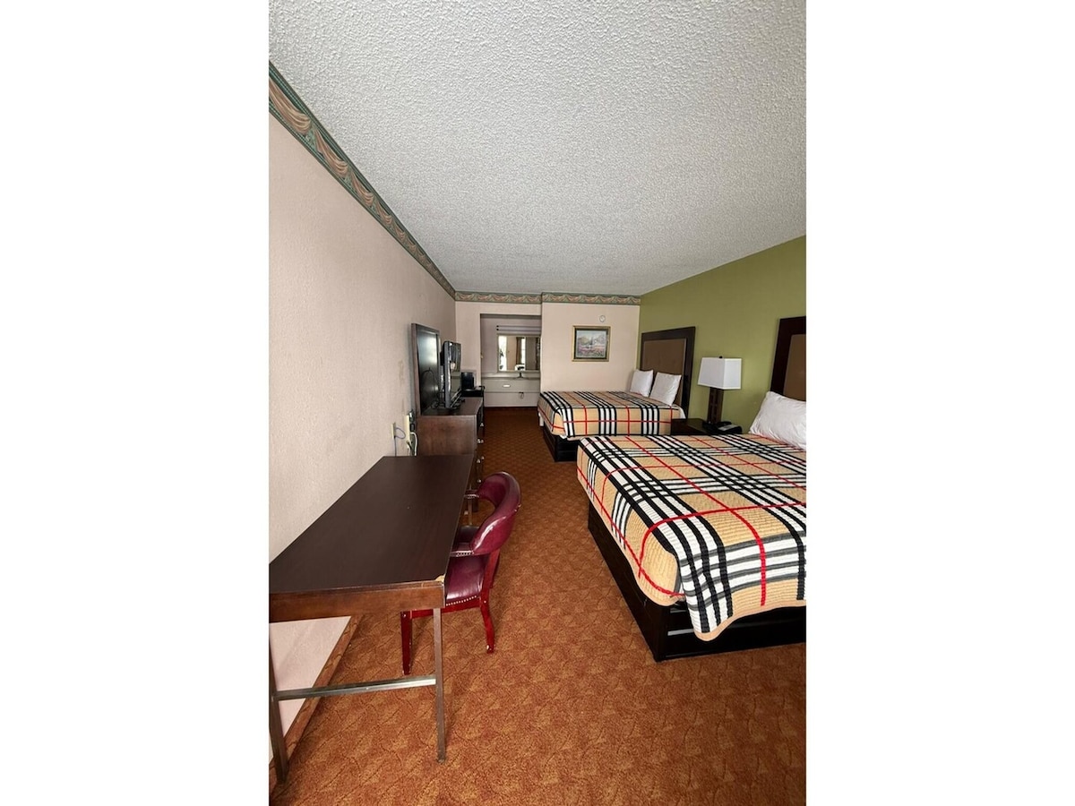 A spacious room is equipped with two queen beds, each adorned with patterned covers. A work desk with a chair is located beside a TV. Warmly colored walls contrast with carpeted flooring, creating a comfortable environment for relaxation.