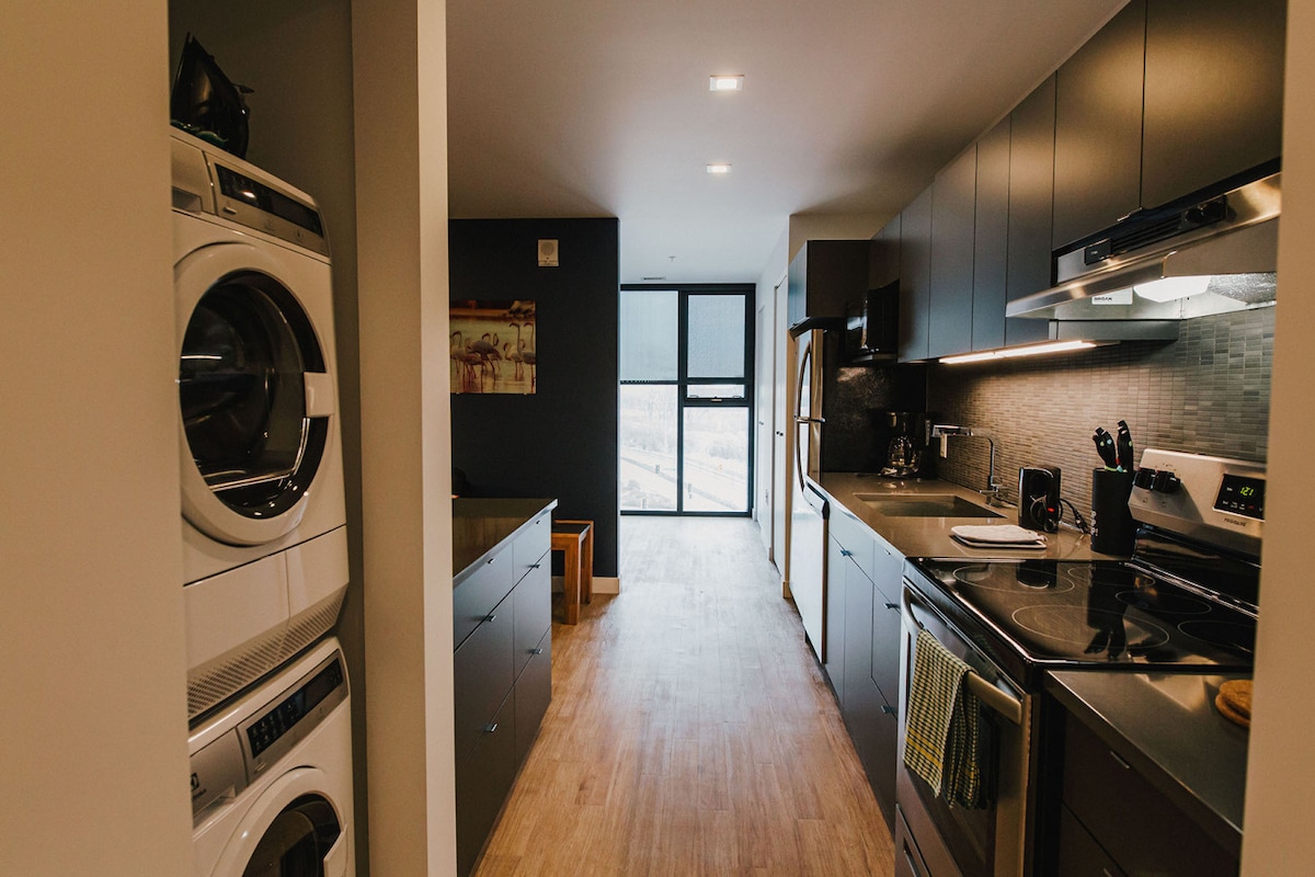 A modern kitchen is featured, showcasing sleek cabinets and countertops. Premium appliances, including a stove and refrigerator, are present. In-suite laundry facilities are integrated into the design. Natural light enters through the floor-to-ceiling windows, enhancing the functional layout.
