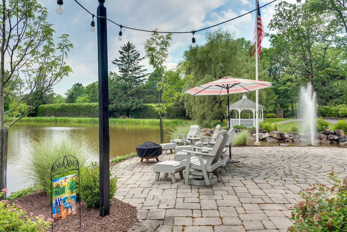 A serene patio area features light-colored Adirondack chairs arranged on a stone surface, overlooking a calm pond. A large umbrella provides shade to one chair, while string lights hang above, creating a pleasant ambiance. A fountain can be seen in the water, adding to the tranquil setting.