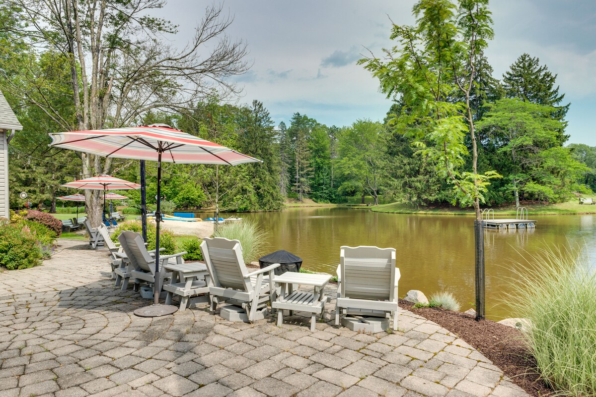 A landscaped patio area features several light-colored lounge chairs and tables under striped umbrellas. The tranquil lake is visible in the background, surrounded by lush greenery and tall trees, creating a serene outdoor setting perfect for relaxation.