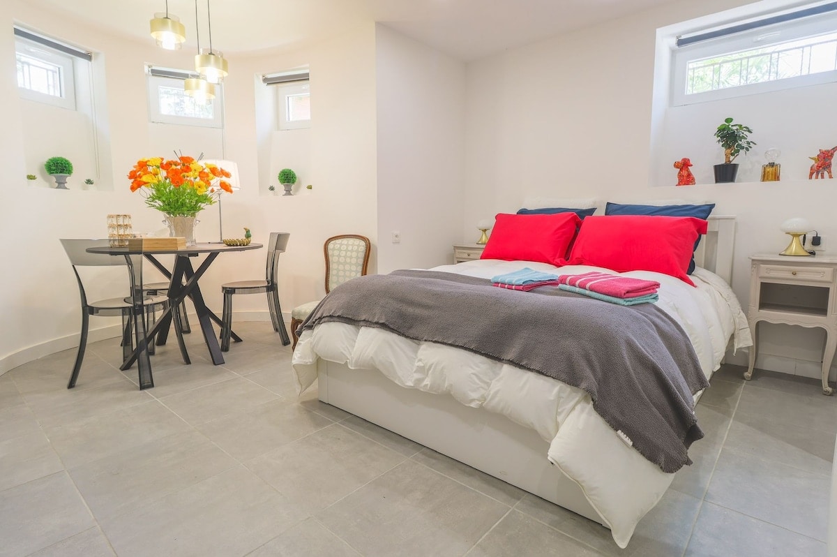 A spacious studio offers a comfortable sleeping area with a large bed dressed in neutral linens and vibrant red pillows. A dining table with transparent chairs is positioned nearby, complemented by decorative plants and a bright floral arrangement. Ample natural light enters through small windows.