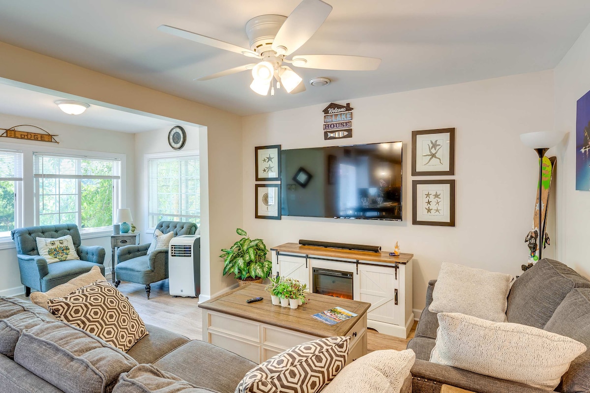 A cozy living room is illuminated by natural light from large windows. A plush couch and love seat are arranged around a coffee table, with a smart TV mounted on the wall. Decorative art frames and plants add warmth to the welcoming space.