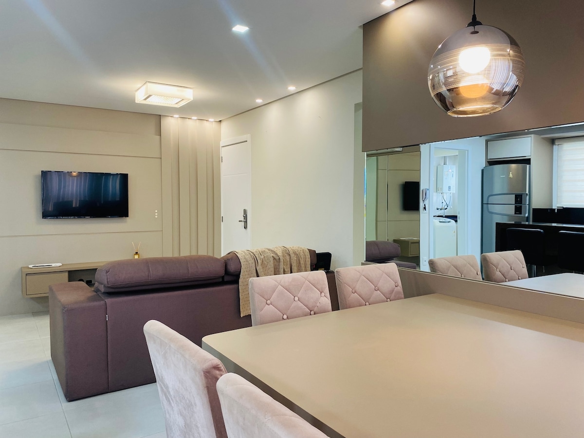The living area features a comfortable seating arrangement with a dark sofa facing a mounted television. A dining table for six is positioned nearby, with elegant chairs adding to the modern aesthetic. Natural light filters through large windows, illuminating the neutral tones of the decor.
