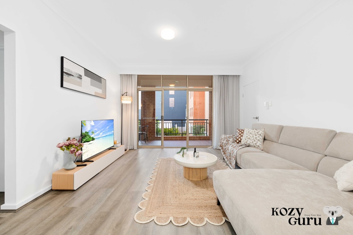 A bright living area featuring a comfortable sofa and a low coffee table on a textured rug. A large flat-screen TV is mounted on the wall with decorative art above it. Glass doors open to a private balcony, allowing natural light to fill the space.