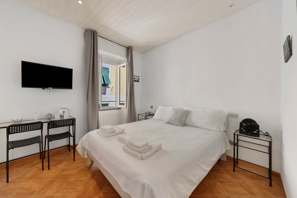A bright and airy bedroom features a spacious bed dressed in white linens, flanked by two modern side tables. A television is mounted on the wall opposite the bed, while natural light filters through a nearby window, accentuated by light curtains. Two chairs and a small table are provided.