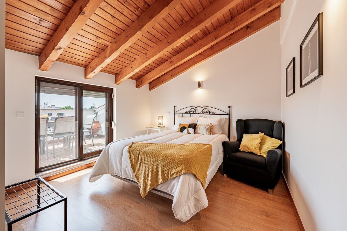 An airy bedroom features a spacious bed adorned with soft linens and a cozy blanket in warm tones. Natural light filters through large glass doors, leading to a balcony. A comfortable armchair complements the room, while wooden beams add texture to the ceiling.