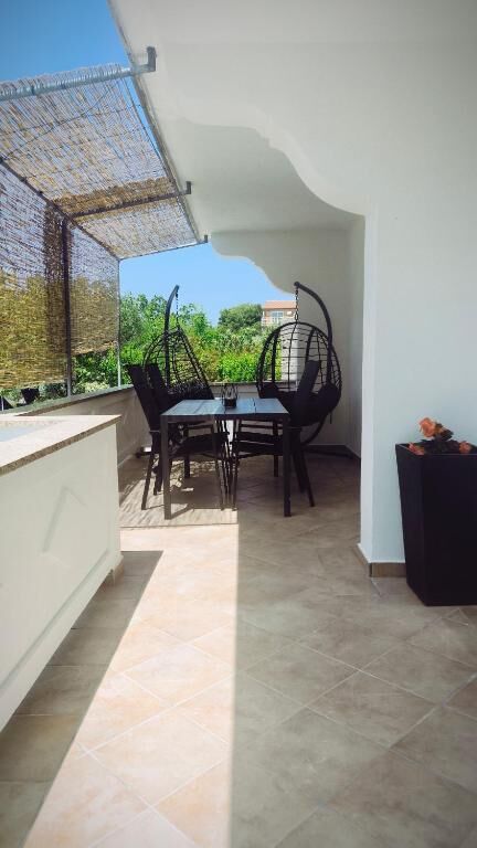 An outdoor seating area is presented, featuring a round table surrounded by four black chairs. Two hanging chairs add a relaxed touch. Sunlight filters through a bamboo shade, creating a play of light on the tiled floor. Greenery is visible in the background.