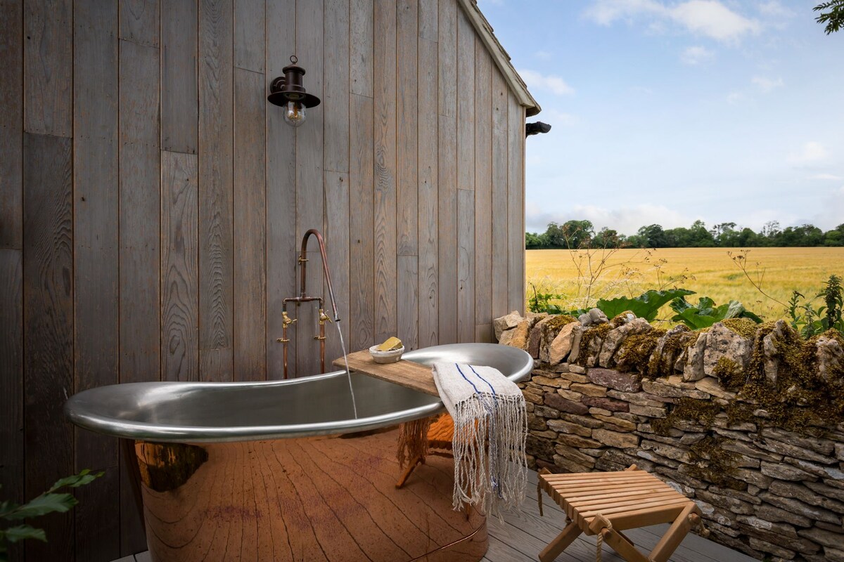 An outdoor copper bath is positioned on a wooden deck, surrounded by rustic wooden walls. A small wooden stool is nearby, while the view beyond features expansive fields and a clear sky, enhancing the serenity of the space.
