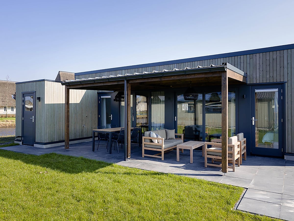 An outdoor terrace is featured with a wooden roof overhang, providing shade. Comfortable seating is arranged around a coffee table, accompanied by additional dining furniture. Lush green grass surrounds the stone-paved area, creating a welcoming space for relaxation and views of the nearby water.