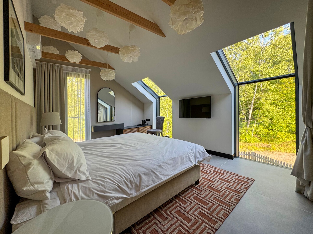 A spacious bedroom is furnished with a comfortable large bed, adorned with white bedding. Large windows provide a view of the surrounding forest, while modern light fixtures hang from the sloped ceiling. A desk and chair are positioned against the wall, enhancing the room's functionality.