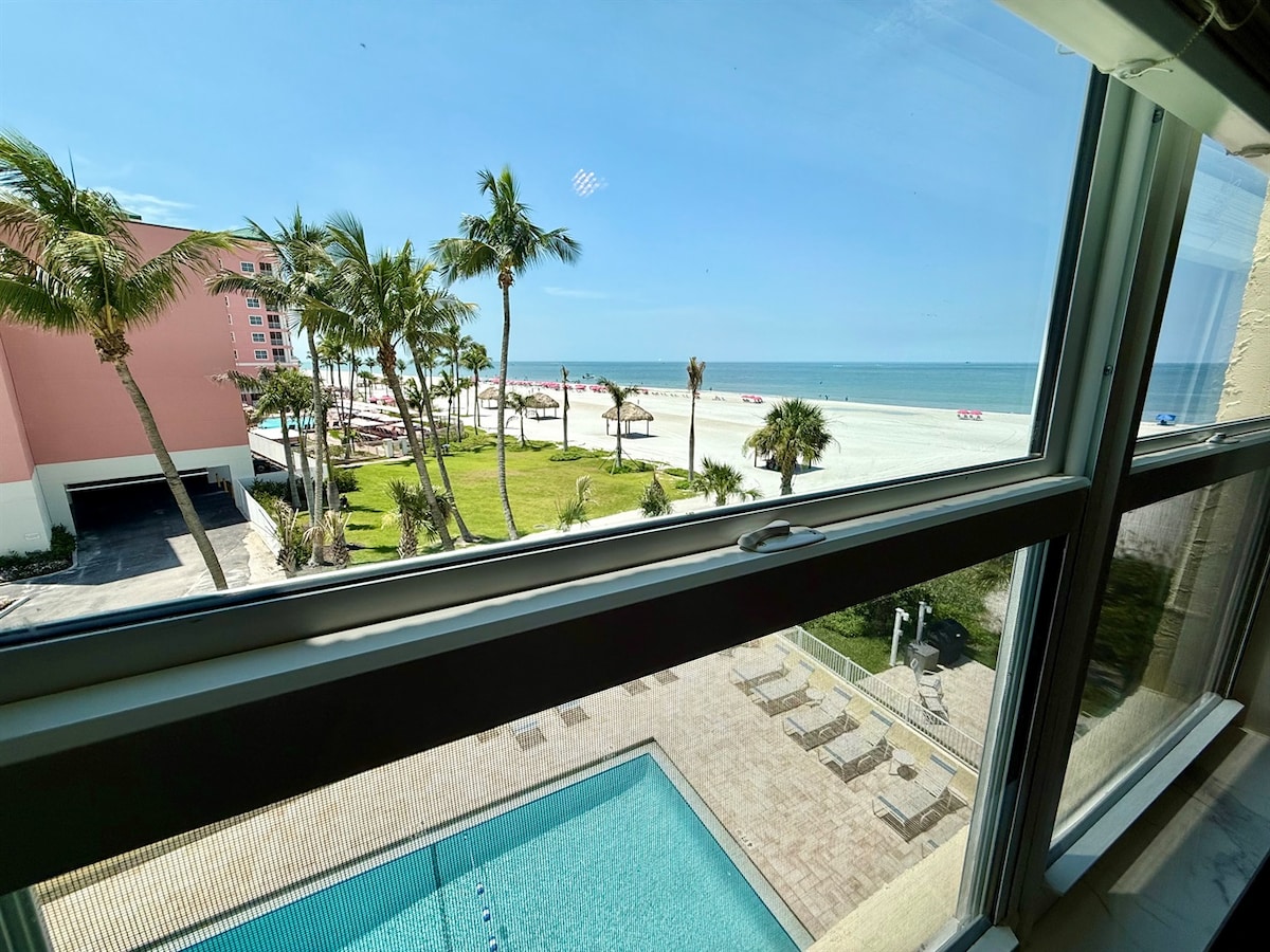 A view from the window highlights the expansive sandy beach meeting the Gulf waters. Palm trees frame the scene, while a beachfront pool area with sun loungers sits below. Sun umbrellas dot the beach, inviting relaxation under clear blue skies.