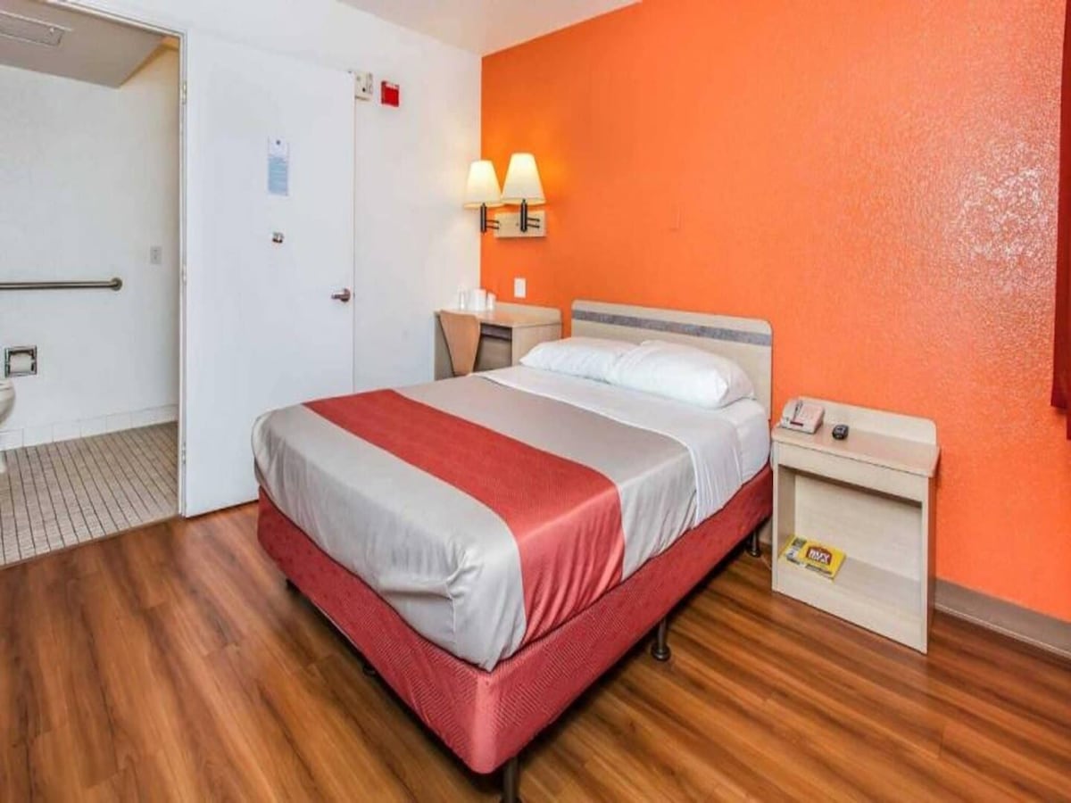 A comfortable room features a spacious bed with smooth linens, placed against a vibrant orange wall. Soft lighting from bedside lamps illuminates the space, and a small table with a telephone is positioned next to the bed. The bathroom area is located in the background.