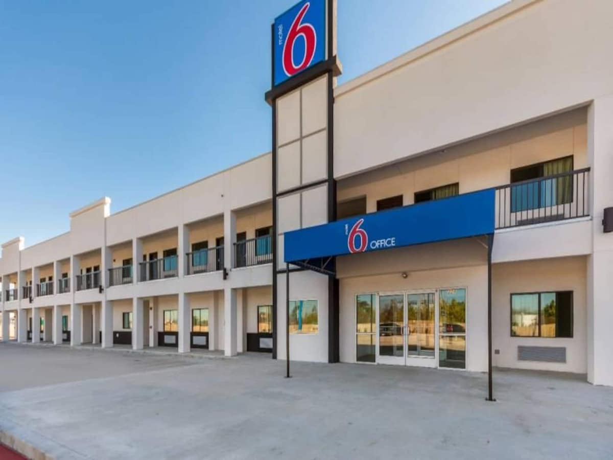 The exterior of the Motel 6 is shown, featuring a simple, modern design with a prominent blue and red sign. Ground-level access is provided through wide glass doors, leading to the reception area. Multiple rooms are visible above, each with balconies and large windows.