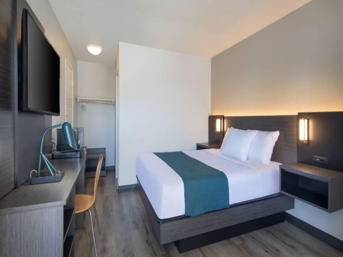 A modern guest room features a spacious bed with crisp white linens and a teal accent blanket. A television is mounted on the wall opposite, and a desk with a chair is positioned nearby. Light wood flooring complements the simple and functional design.
