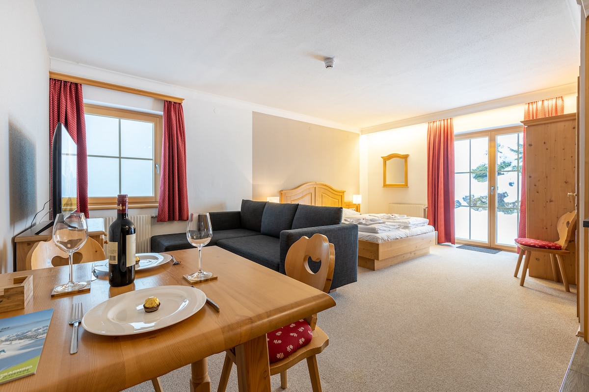 A spacious studio apartment is shown, featuring a comfortable seating area with a dark sofa and a dining table set for two. A double bed is partially visible, positioned against a light wood frame. Large windows allow natural light, with light curtains framing the views. A light carpet covers the floor.