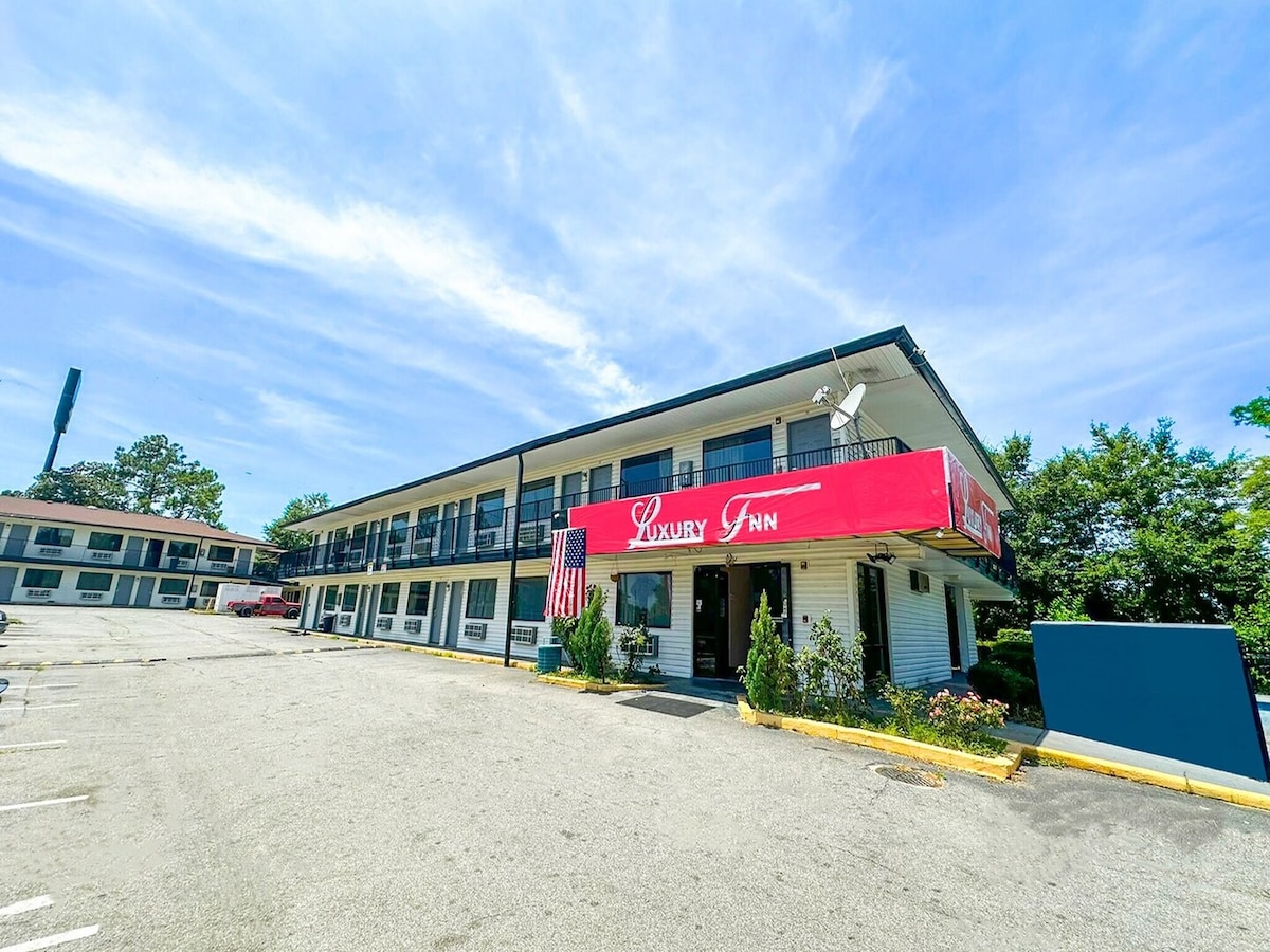 The exterior of the hotel exhibits a two-story building with a red awning labeled 'Luxury Inn.' Surrounded by well-maintained landscaping, the parking area features spaces for guests. A clear blue sky enhances the overall appearance of the property.