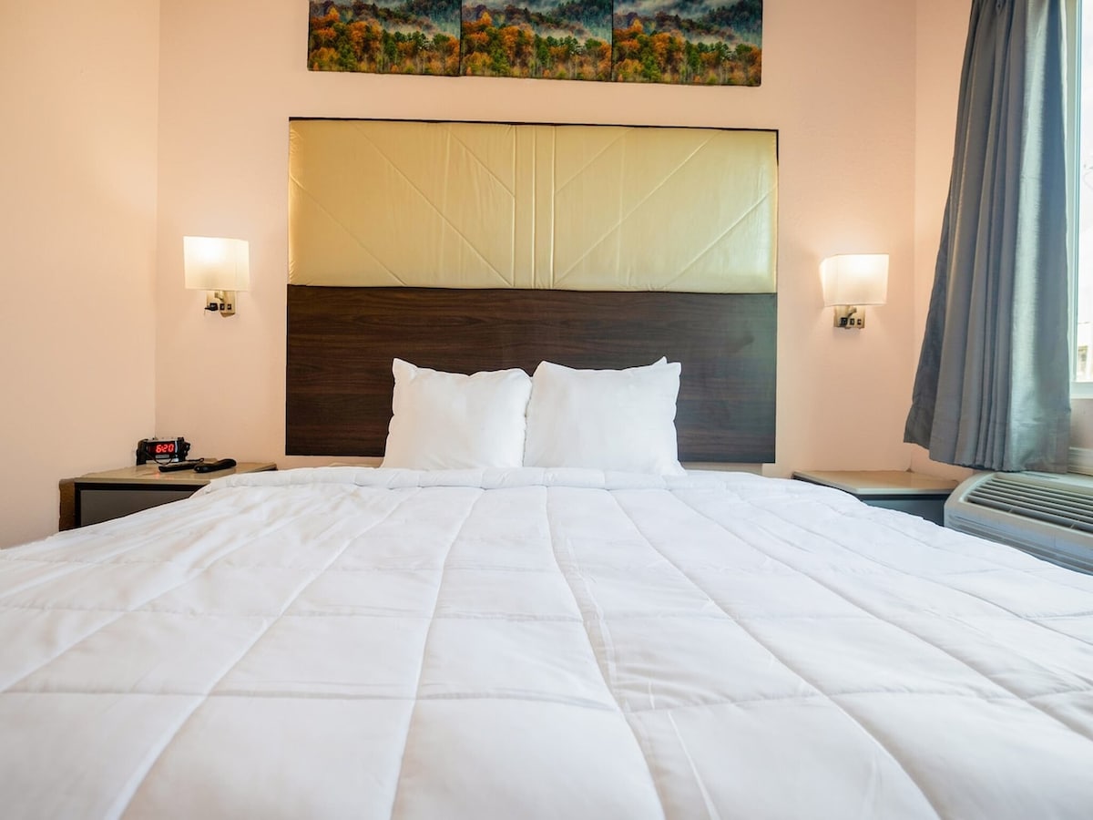A spacious bed with white bedding is centered against a wall featuring a padded headboard. Soft lighting emanates from two bedside lamps, complementing a window with gray curtains. A wooden nightstand holds a clock and an air conditioning unit is visible in the background.