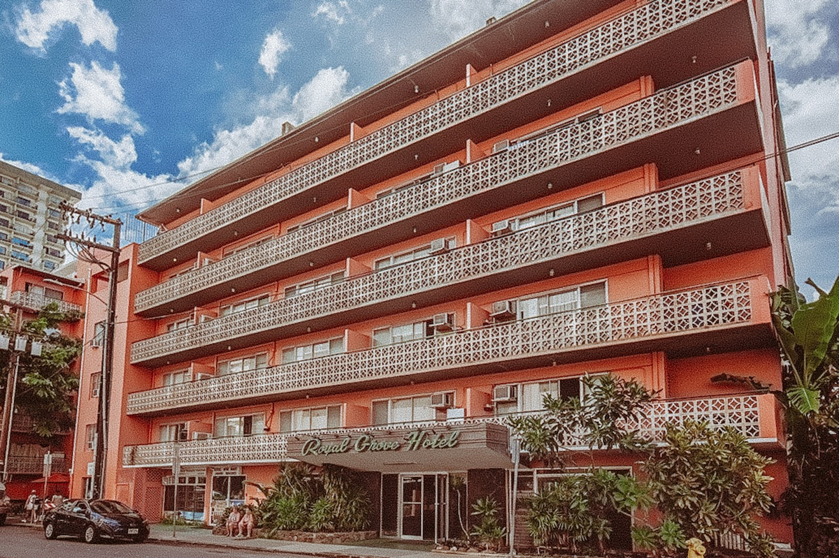 The exterior of the Royal Grove Hotel is depicted, showcasing a multi-story building characterized by pink walls and decorative balconies. Green foliage surrounds the entrance, while multiple air conditioning units are visible on the building's facade. A clear blue sky with scattered clouds forms the backdrop.