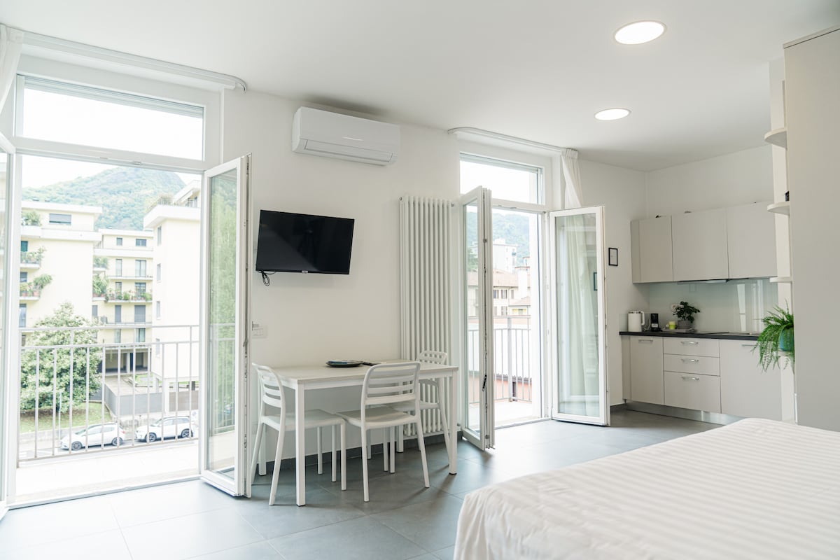 A modern studio space features large, open windows that invite natural light. A small dining table with white chairs is situated next to a wall-mounted television, while an equipped kitchenette is visible in the background. Light-colored cabinetry complements the airy feel of the room.