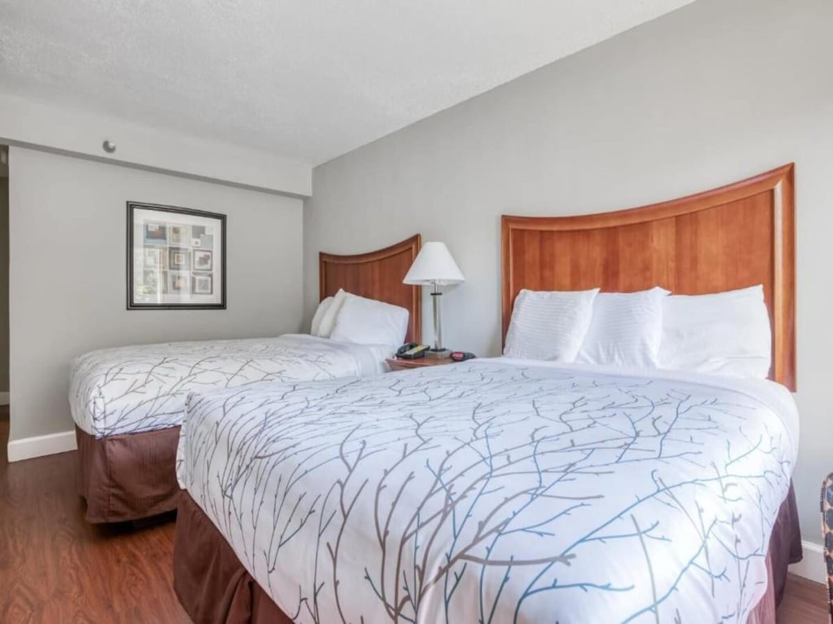 A guest room features two spacious queen beds with light-colored bedding, complemented by wooden headboards. A bedside lamp rests on a small table between the beds. A framed artwork hangs on the wall, enhancing the room's calming atmosphere.