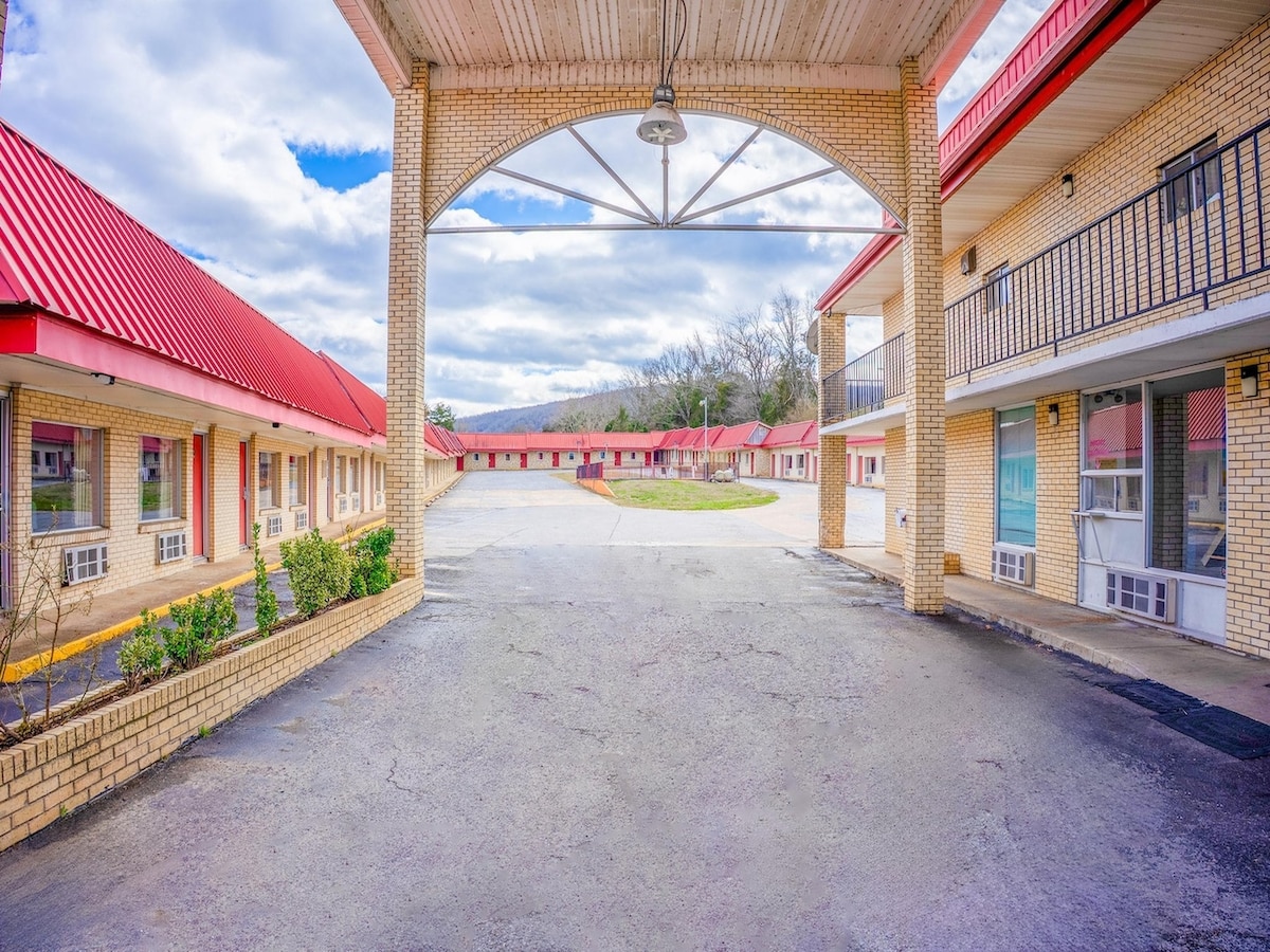 A view of the hotel exterior features a central courtyard, with guest rooms arranged around a paved area. The red rooftops contrast against the light brick facade. A covered archway provides entry, and the landscaped borders add a touch of greenery.