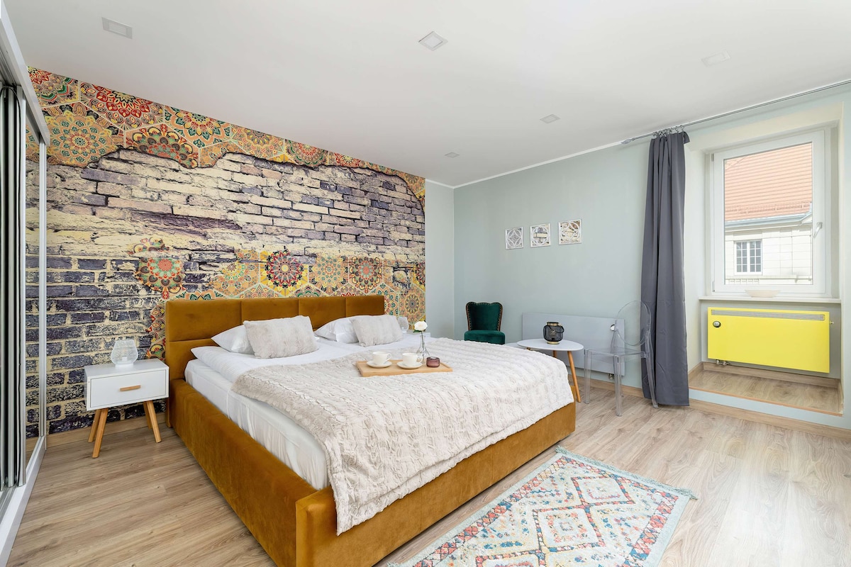 An intimate studio room features a large double bed with a soft light blanket and decorative pillows. A colorful accent wall showcases brick patterns, while natural light enters through the window. A small seating area with a green chair and a table adds functionality to the space.