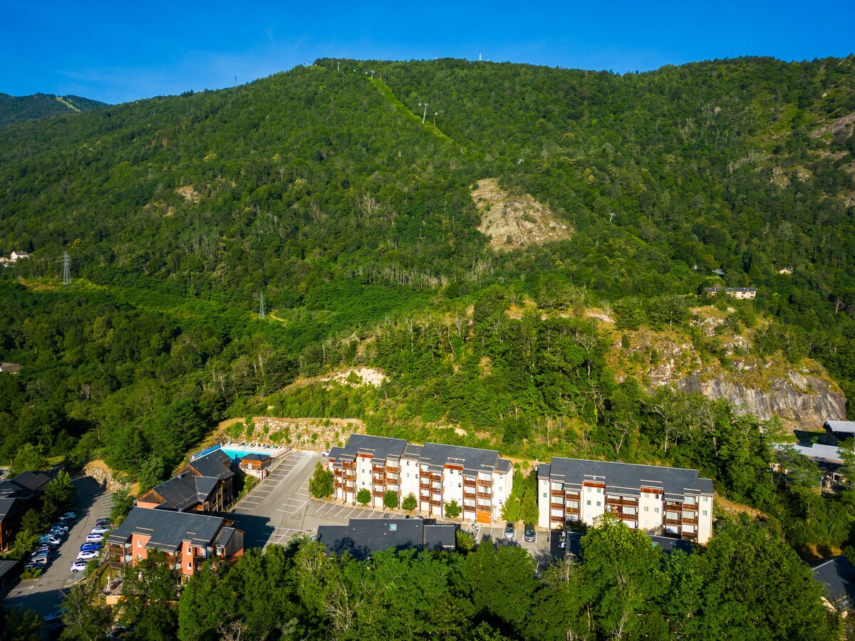 An aerial view captures the expansive greenery of the surrounding mountains and the structured layout of accommodation buildings. An outdoor swimming pool is visible on-site, complemented by ample parking space and pathways amid the natural landscape.