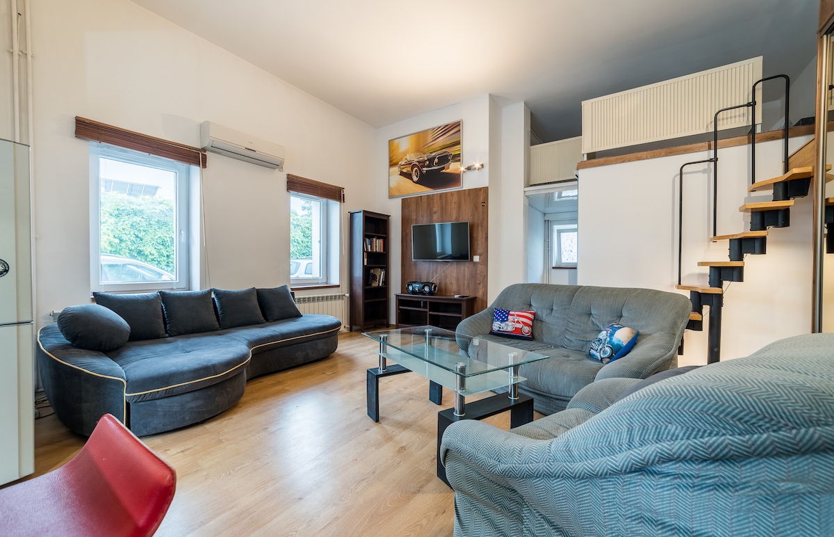 The living room features comfortable seating arrangements with a curved sofa and a separate armchair. A glass coffee table is positioned at the center, complemented by a TV mounted on the wall. Stairs lead to an upper loft area, adding vertical space to the room.