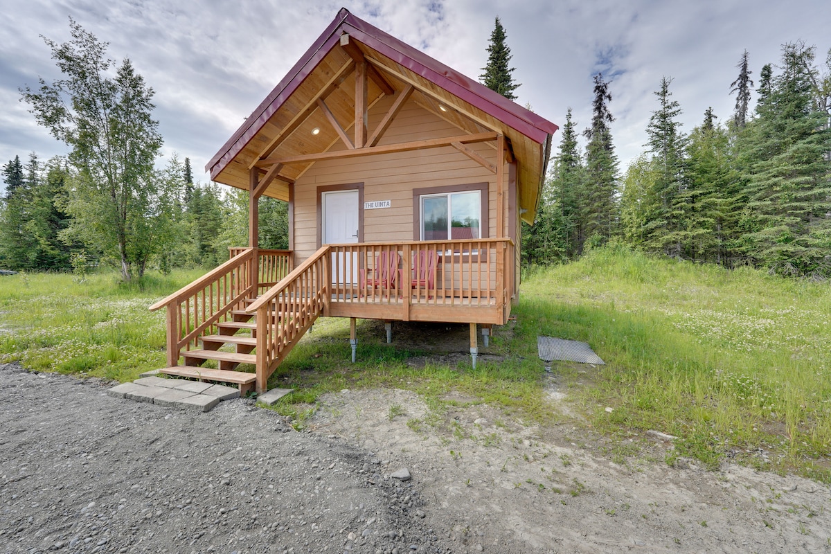 A single-story cabin is visible with a wooden exterior and a red roof. A front porch with a railing provides access, leading to a pathway lined with gravel. Surrounding greenery includes trees and wild grass, contributing to the natural setting.