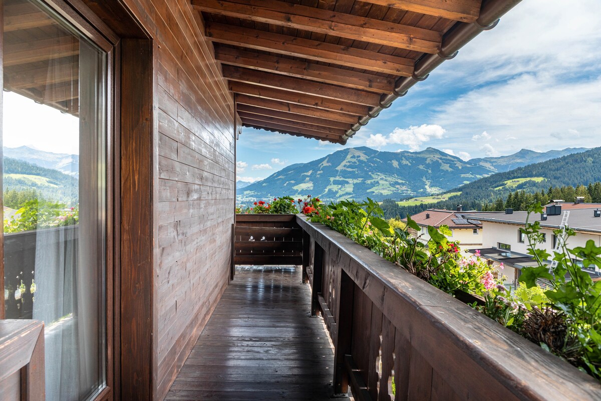 A wooden balcony extends outward, bordered by greenery and colorful flowers. The breathtaking view showcases rolling hills and alpine mountains, with a clear blue sky above. The balcony's rustic design features slanted wood beams and a warm wooden railing, creating a tranquil outdoor space.